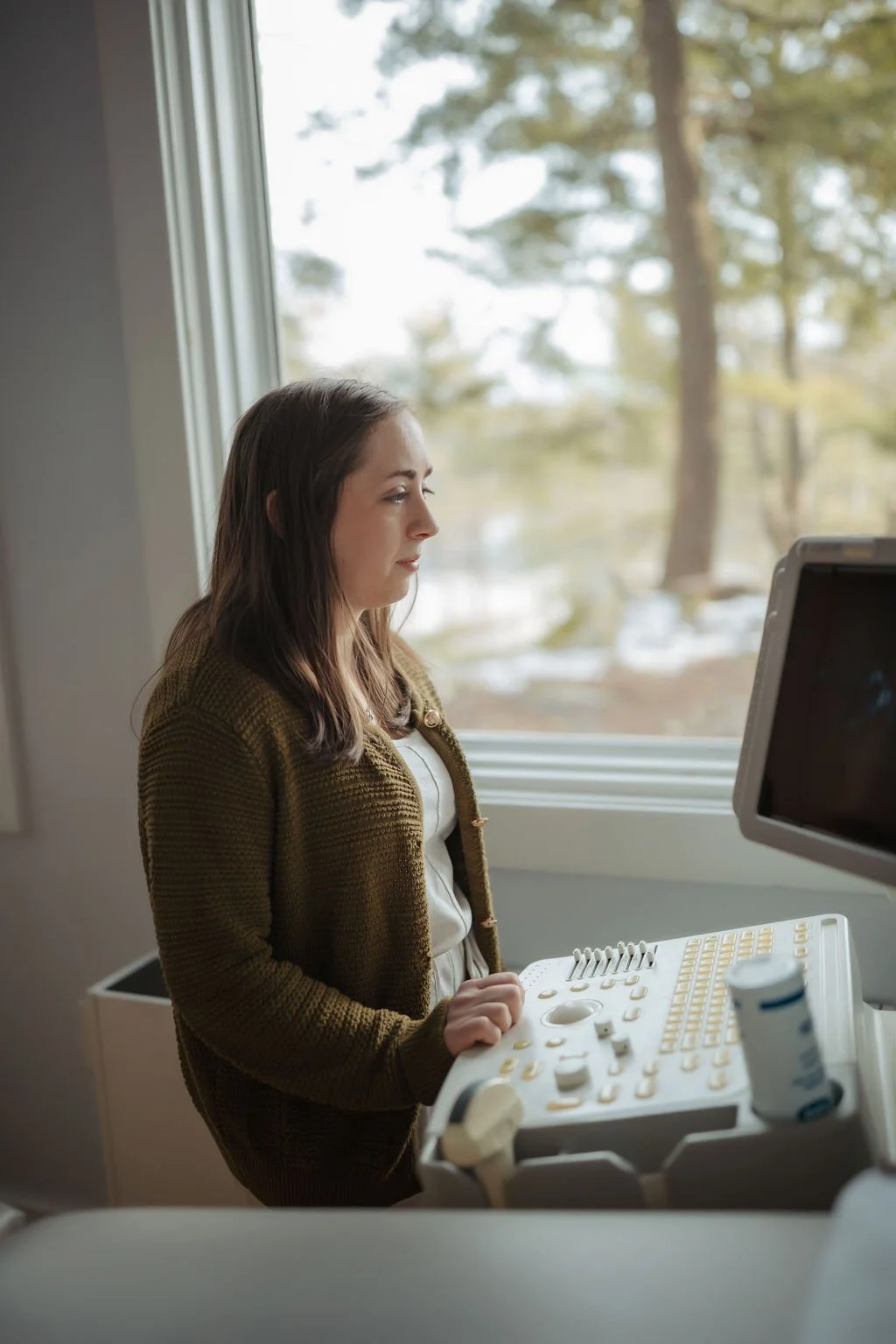 A woman in a brown cardigan operating an ultrasound machine in a medical office, with a window showing trees outside.
