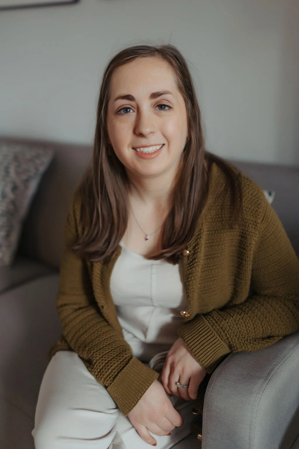 A smiling woman with shoulder-length brown hair, wearing a white top and a brown cardigan, sitting on a gray sofa in a living room.