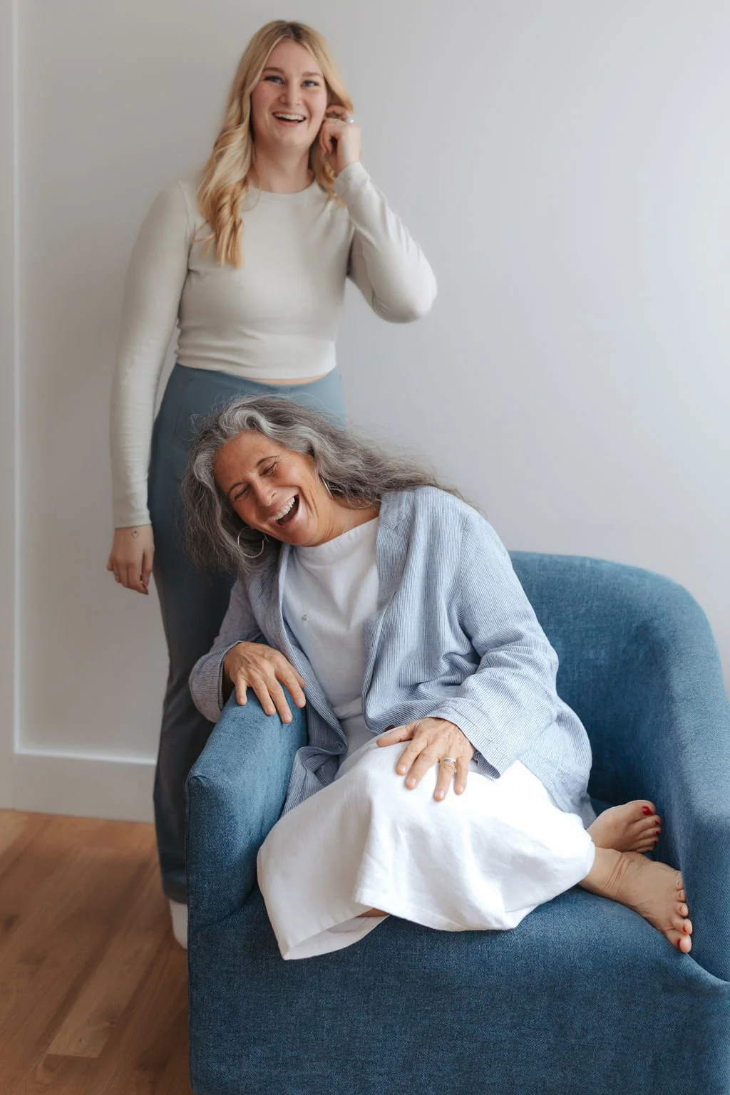 Two women laughing and enjoying each other's company; one is standing and smiling, the other is sitting on a blue armchair with a gray sweater and a white dress, laughing with her eyes closed.