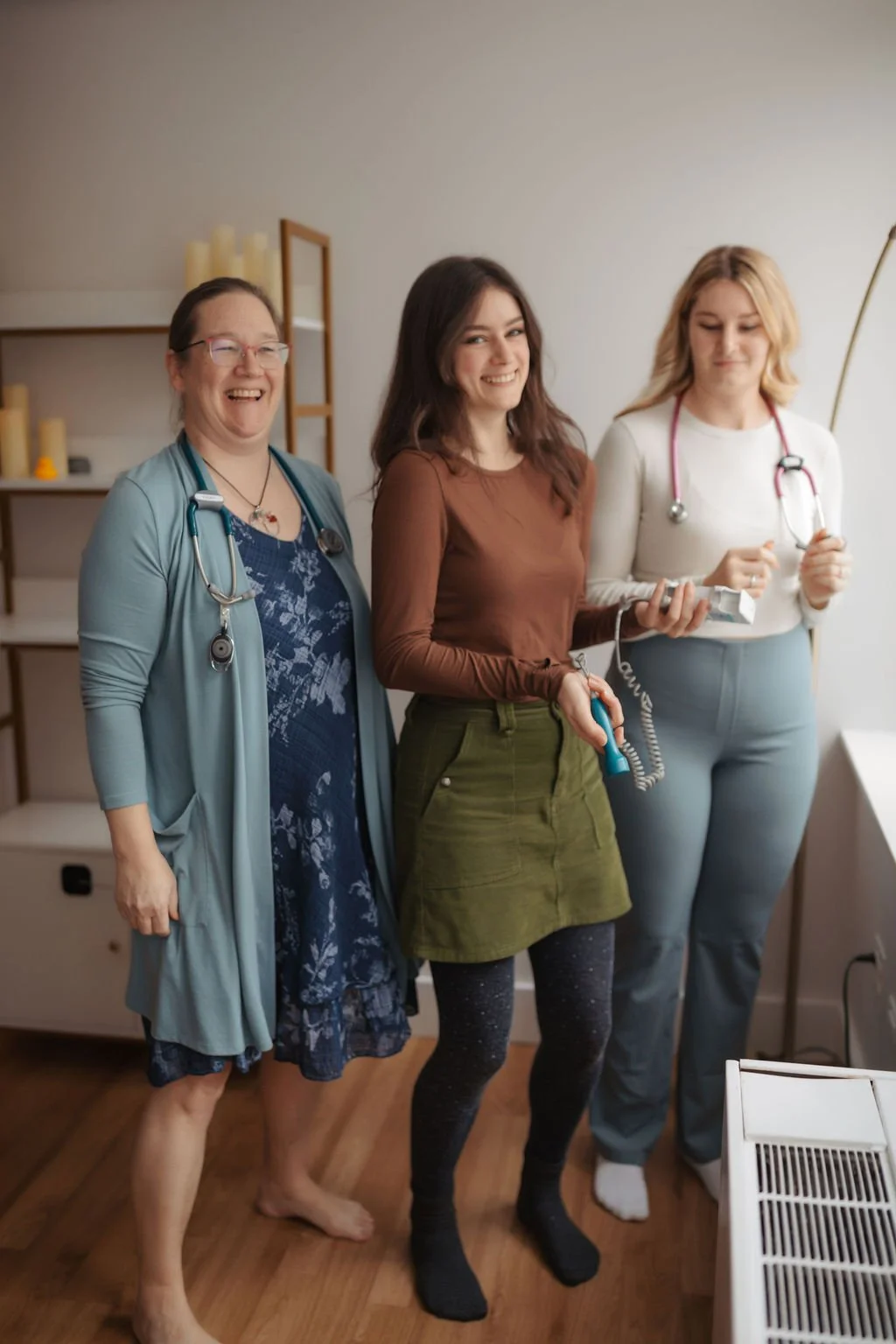 Three women in medical attire, with stethoscopes, smiling and posing in a medical office.
