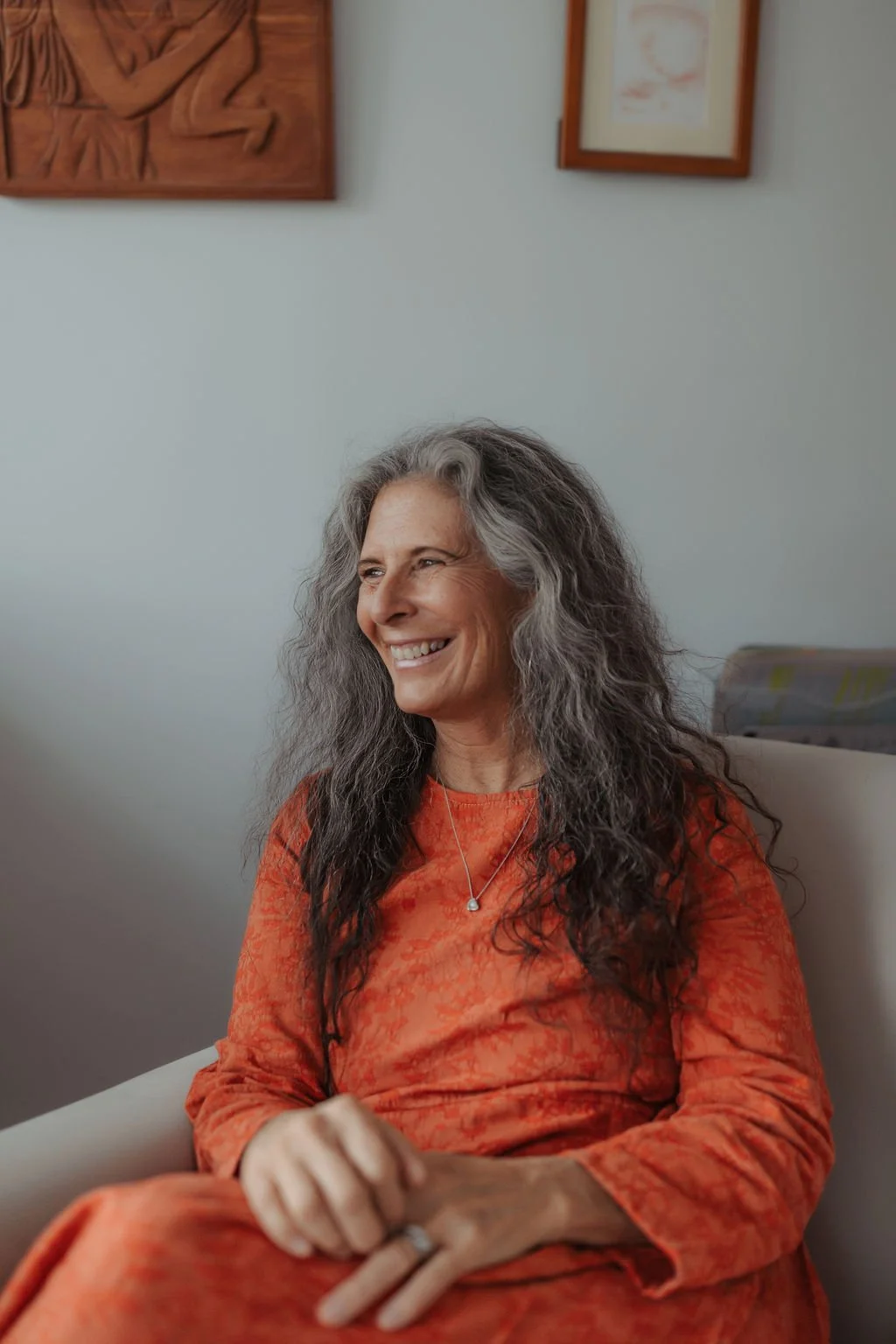 A smiling woman with gray, curly hair sitting indoors against a light gray wall with framed artwork. She is wearing an orange patterned dress and a necklace with a small pendant.