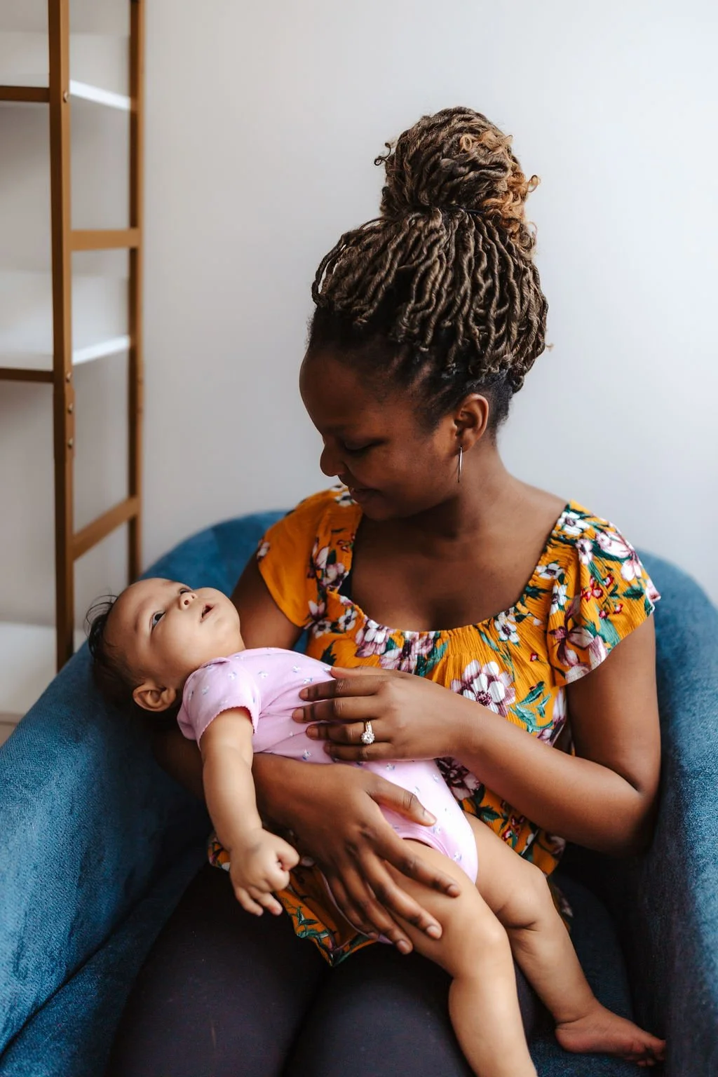 A woman with styled dreadlocks in a high bun holding a baby girl in her arms on a blue couch against a plain white wall.