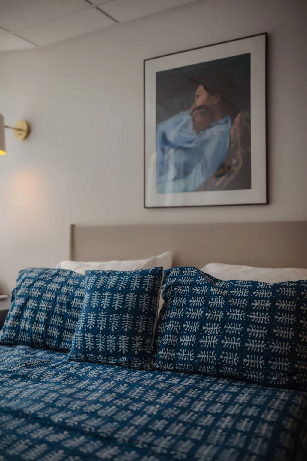 A hotel room bed with blue patterned bedding and pillows, a beige headboard, a wall lamp, a framed painting of a woman, and a plain white wall.