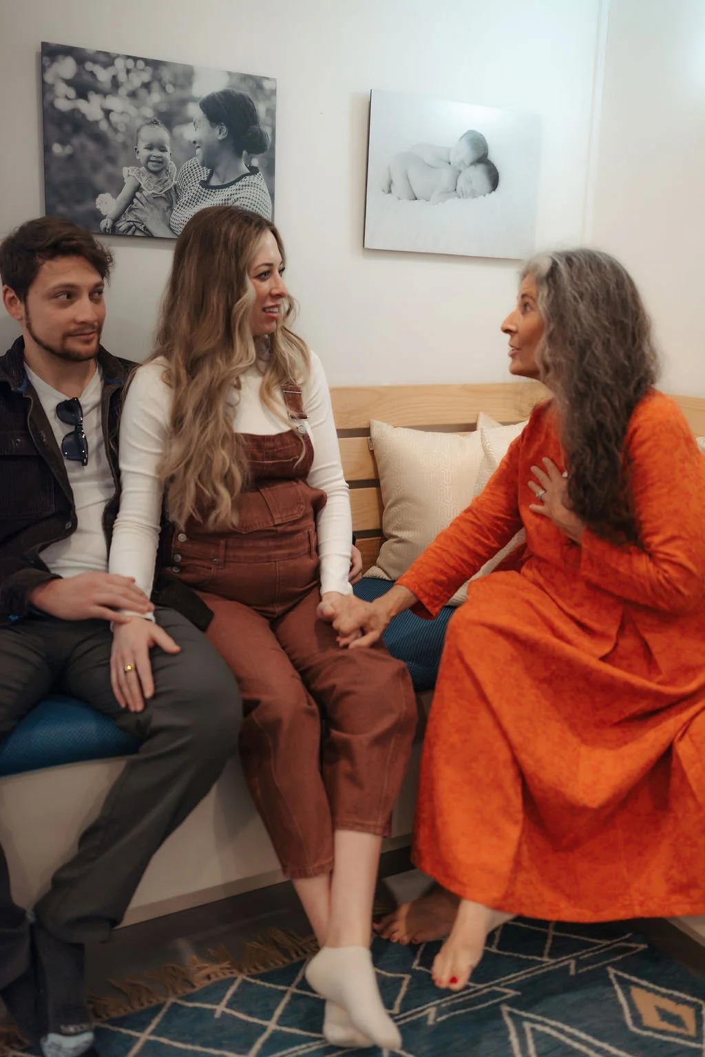 Three people sitting on a couch in a living room, engaging in a conversation. An older woman in an orange dress is touching the hand of a young woman with long, wavy hair, who is wearing a white long-sleeve shirt and brown overalls. A young man in a 