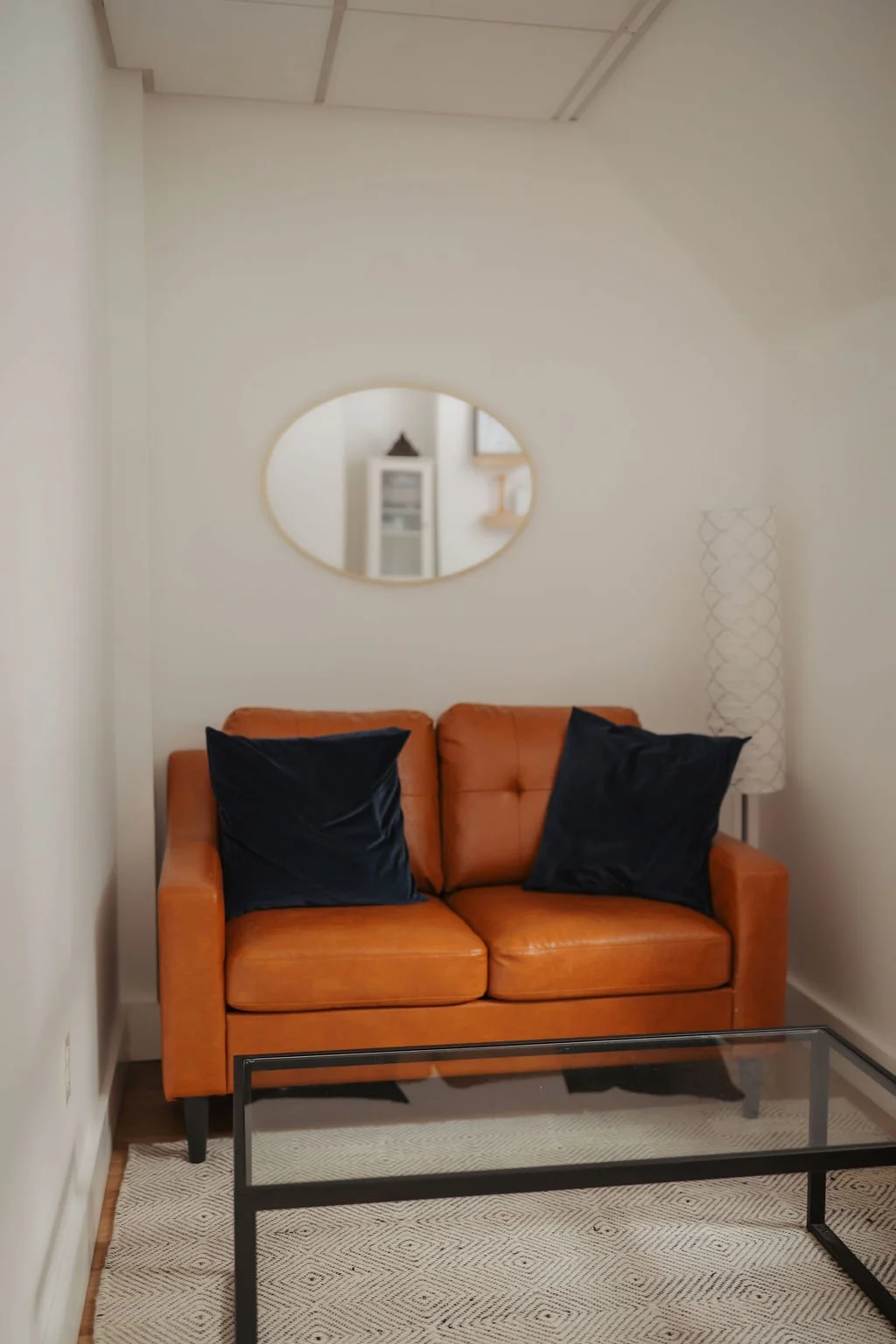 A living room corner with an orange leather sofa, two black pillows, a round mirror on the wall, and a white floor lamp to the right.
