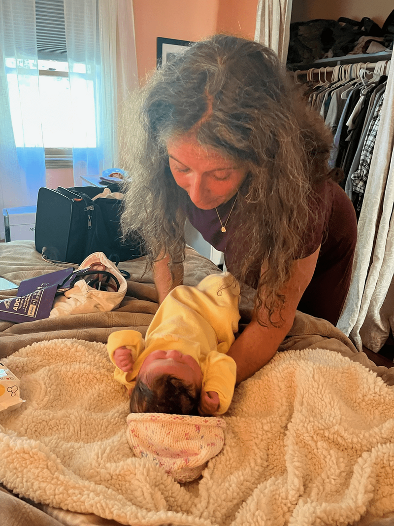 A woman with long curly hair holds a baby dressed in a yellow outfit on a fluffy blanket in a cozy bedroom.
