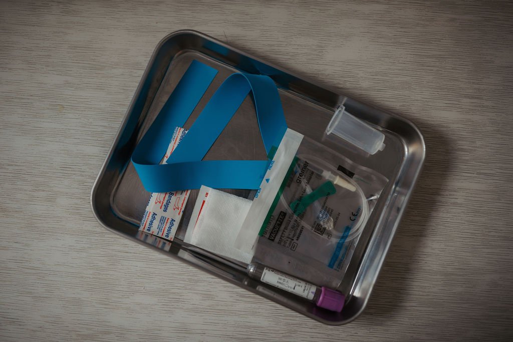 Medical supplies inside a stainless steel tray, including a blue tourniquet, bandages, gauze, a syringe, and a vial, on a wooden surface.