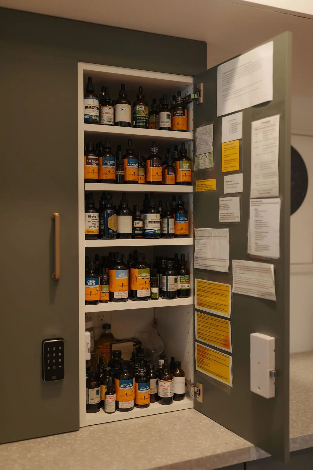 Open cabinet filled with various bottles of essential oils and herbal tinctures, with paper notices and labels attached to the inside of the door.