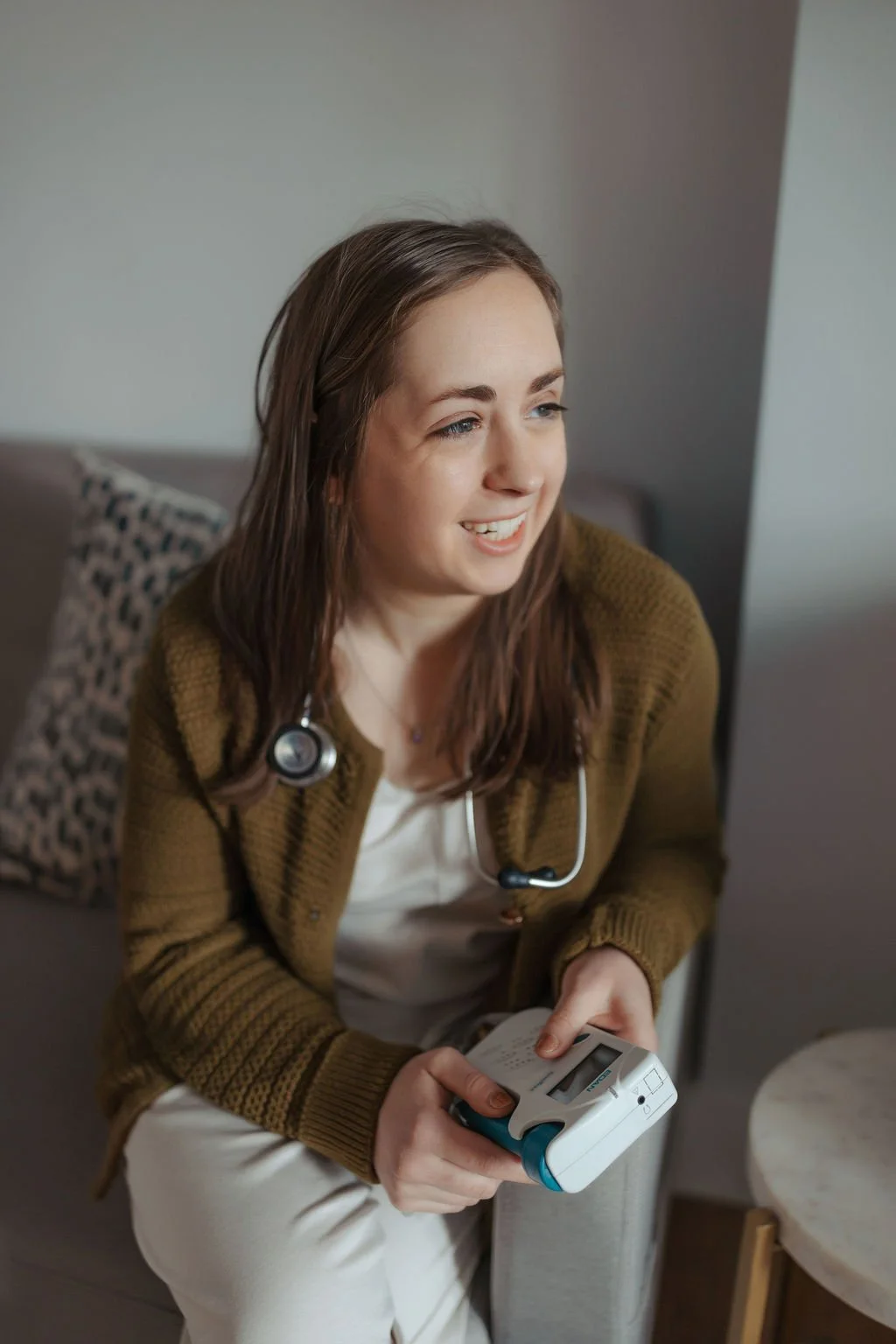 A young woman with brown hair, wearing a brown sweater and white top, sitting on a couch with a leopard print pillow behind her. She is smiling and holding a portable medical device, with a stethoscope around her neck.