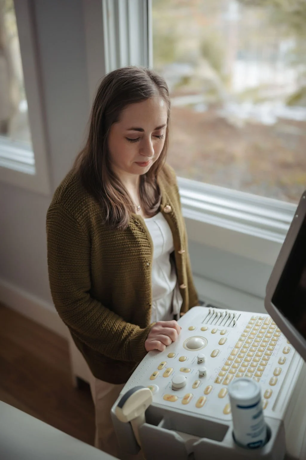 A woman operating an ultrasound machine in a medical setting, with a window and blurred outdoor scene in the background.