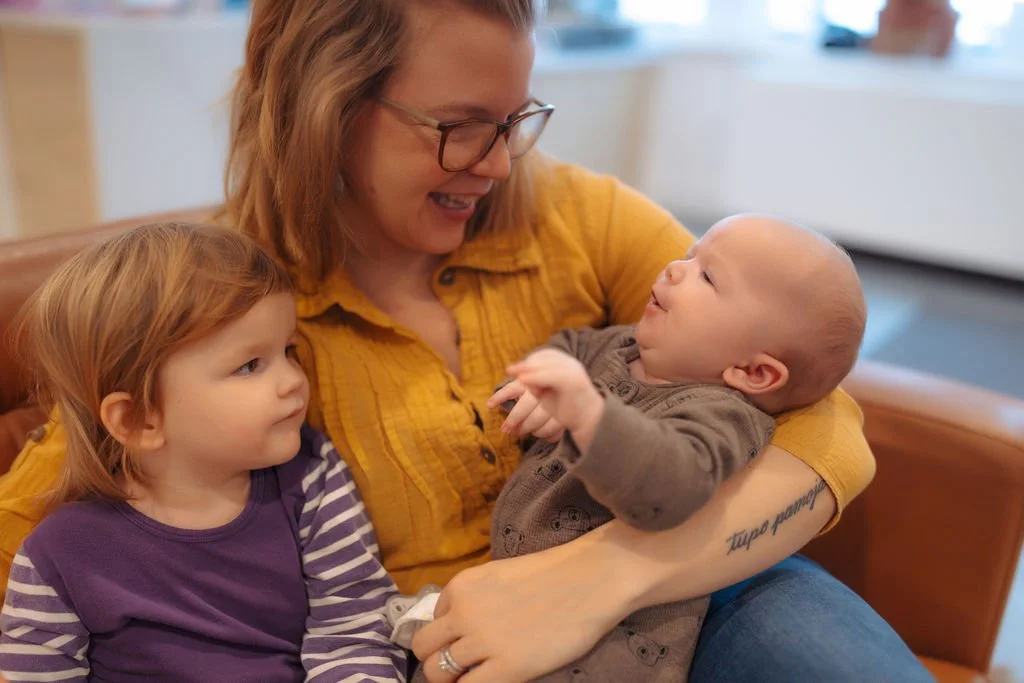 A woman with glasses and a yellow shirt holding a baby boy and looking at him with a smile, while a young girl with red hair and a purple striped shirt looks on.