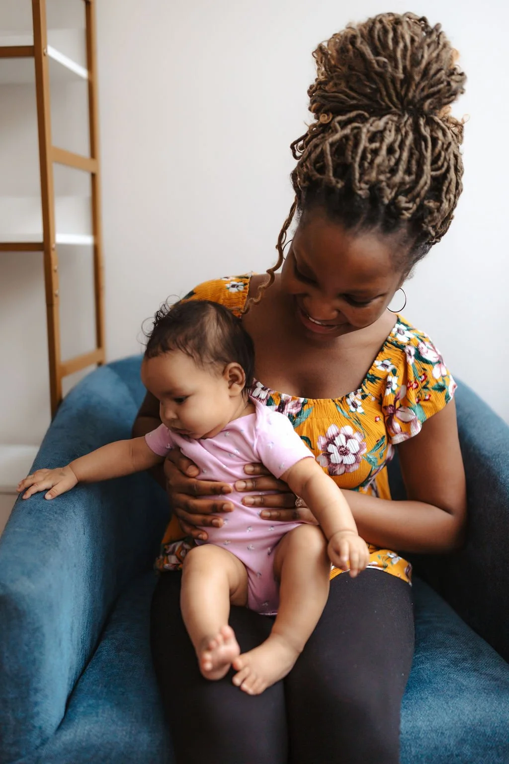 A woman with braided hair and a floral dress sitting on a blue couch, holding a baby dressed in pink. The woman is smiling as she looks down at the baby.