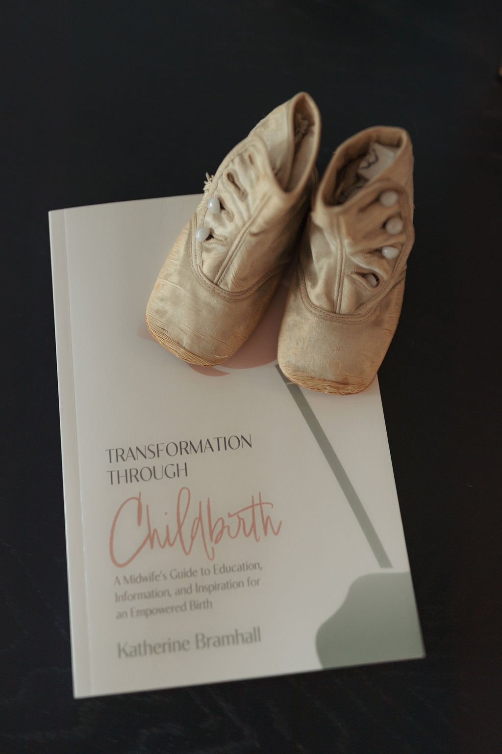 A pair of worn ballet slippers placed on top of a book titled 'Transformation Through Childbirth' by Katherine Bramhall.