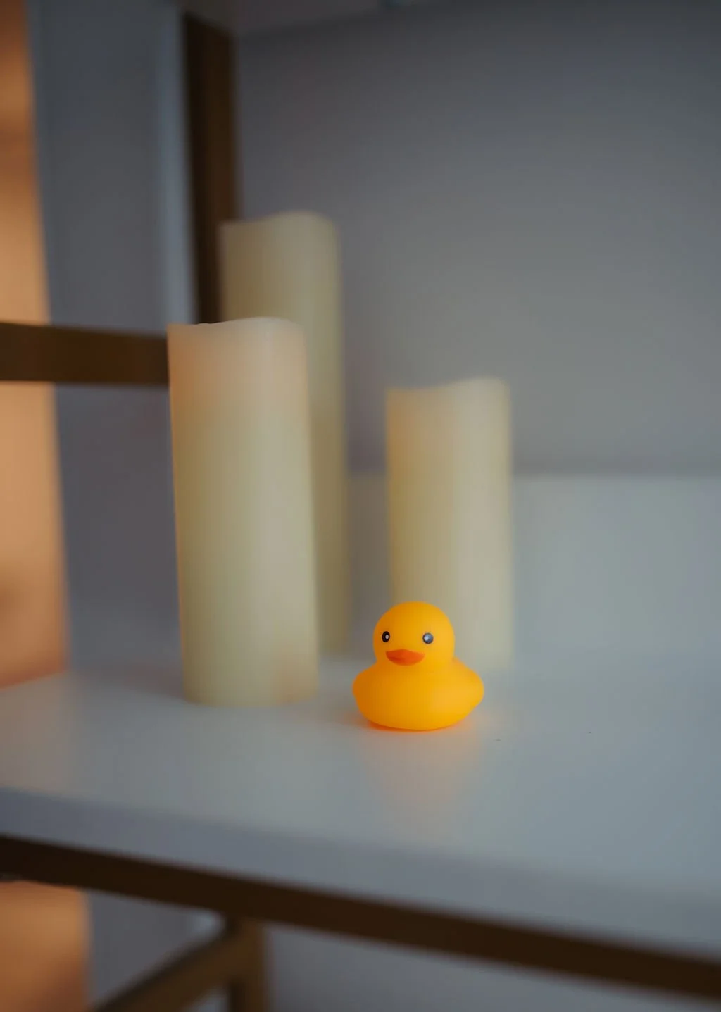 A small yellow rubber duck with a surprised expression on its face, sitting on a white surface in front of three tall, ivory-colored candles, with a wooden shelf partly visible on the left.
