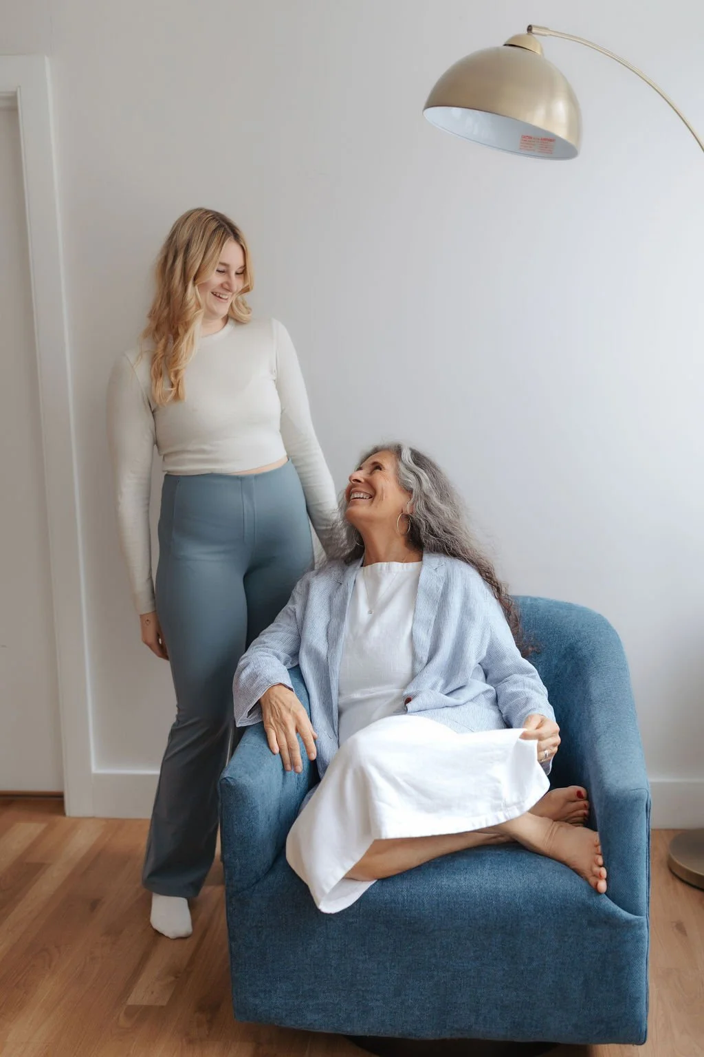 A young woman standing and smiling at an elderly woman sitting on a blue armchair, both appearing happy and engaged in conversation in a bright room.