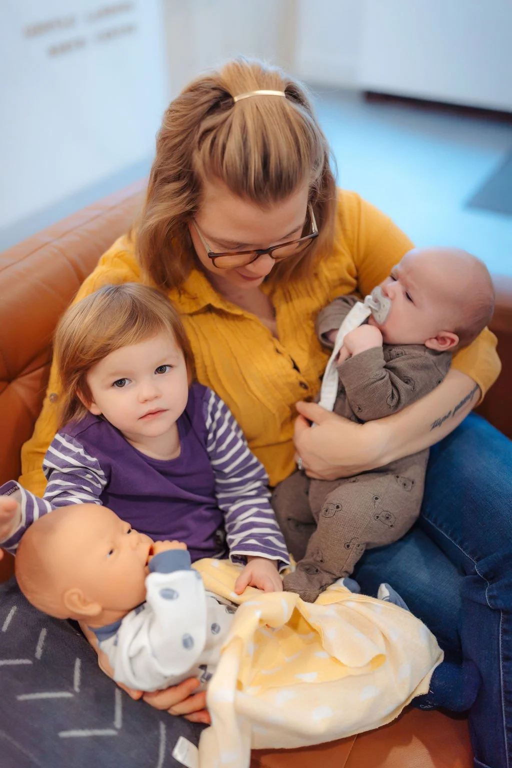 A woman with glasses and light brown hair, wearing a yellow top, sitting on a brown couch with four young children. One child is breastfeeding, another with red hair and a purple top, and two babies, one with a pacifier and the other grasping a bottl