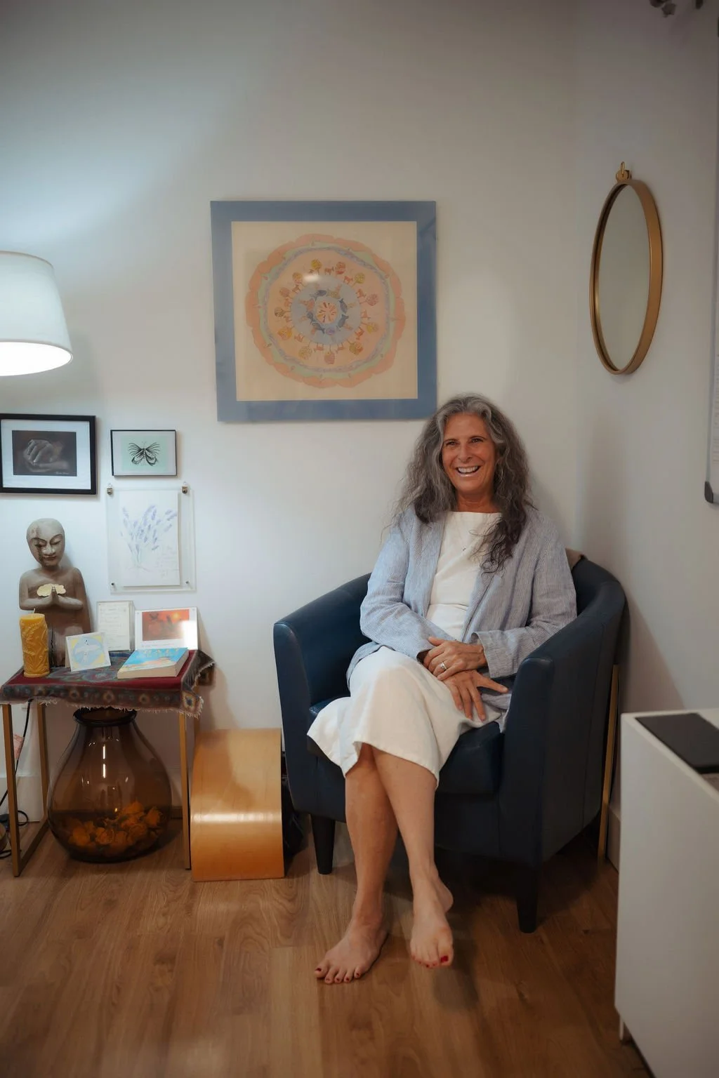 A woman with gray hair sitting in a black armchair, smiling, in a room decorated with artworks and artifacts on the wall and a side table with items on it.