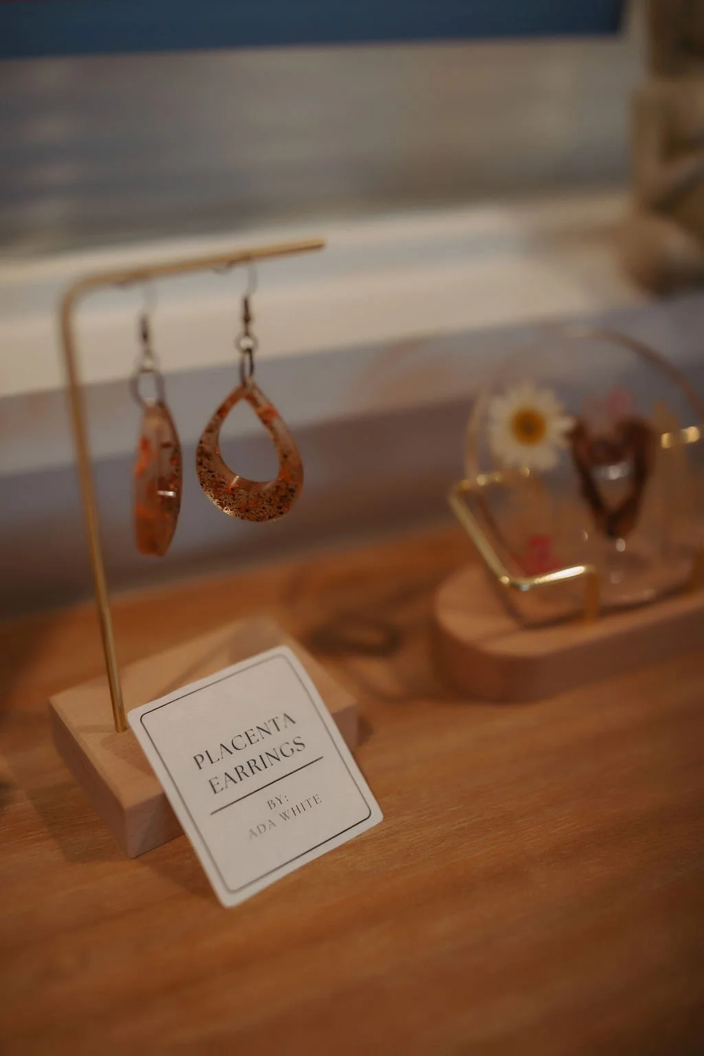 Rust-colored earrings hanging on a display stand at a jewelry shop, with a sign reading 'Placenta Earrings by Ada White' placed on a wooden surface.