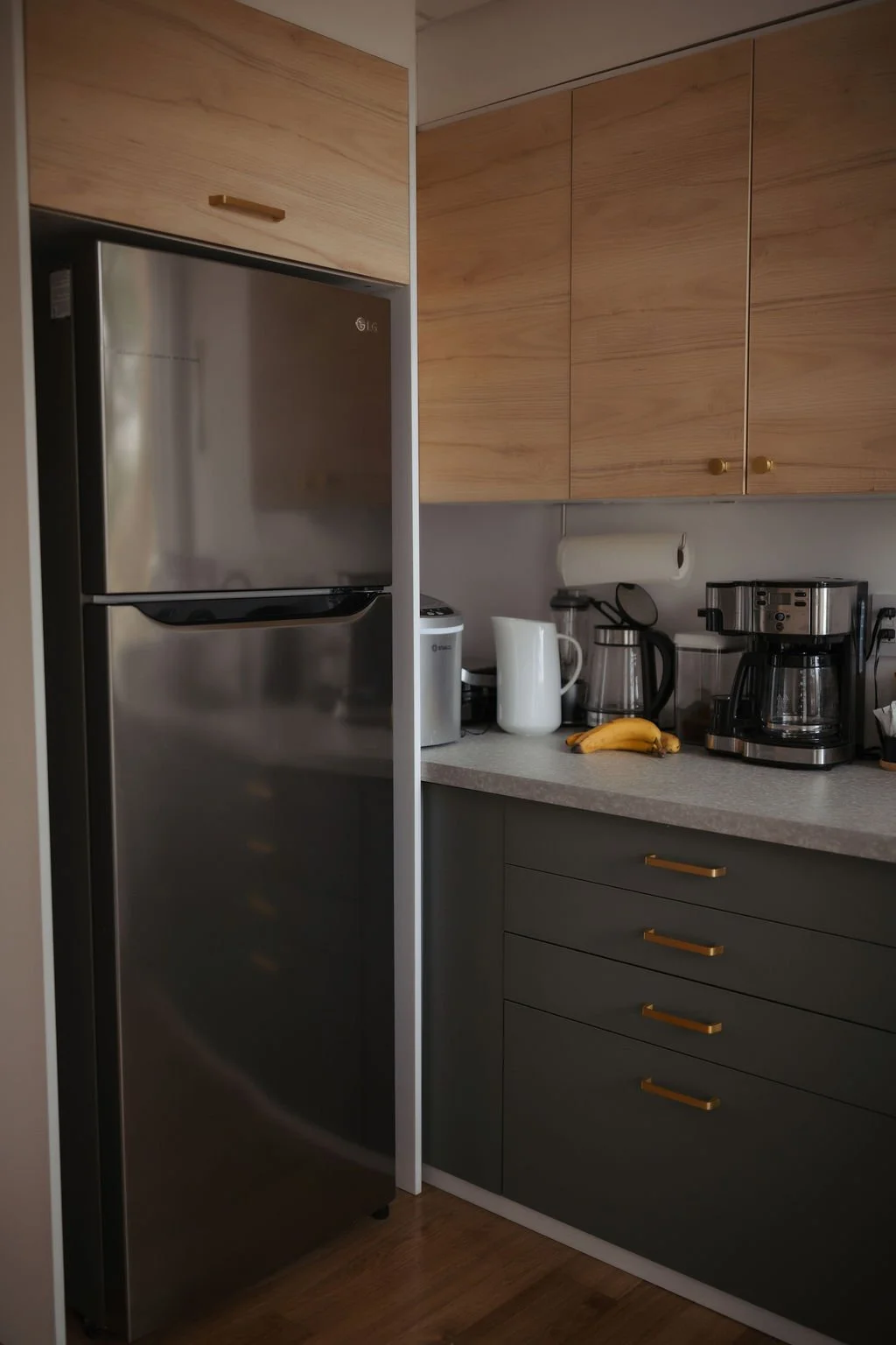 Kitchen with stainless steel refrigerator, light wood cabinets, coffee maker, bananas, paper towel, white pitcher, kettle, and other small appliances on the countertop.
