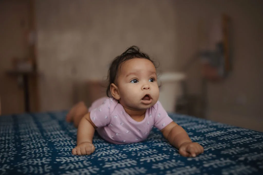 A baby girl lying on her stomach on a bed, looking surprised or curious.