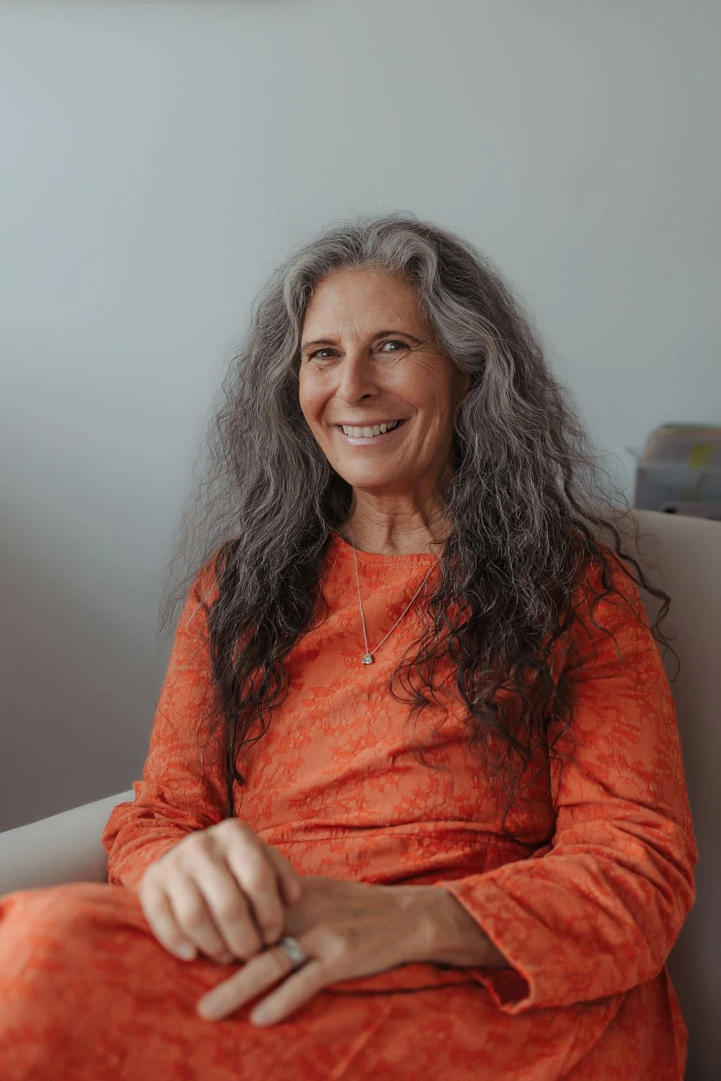 A smiling senior woman with long gray hair, wearing an orange patterned dress and a silver necklace, sitting on a beige chair in an office environment.