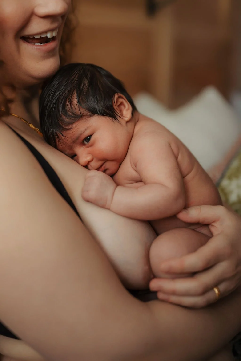 A woman holding a newborn baby close to her chest, smiling with her mouth open while the baby looks relaxed and comfortable, resting on her shoulder.