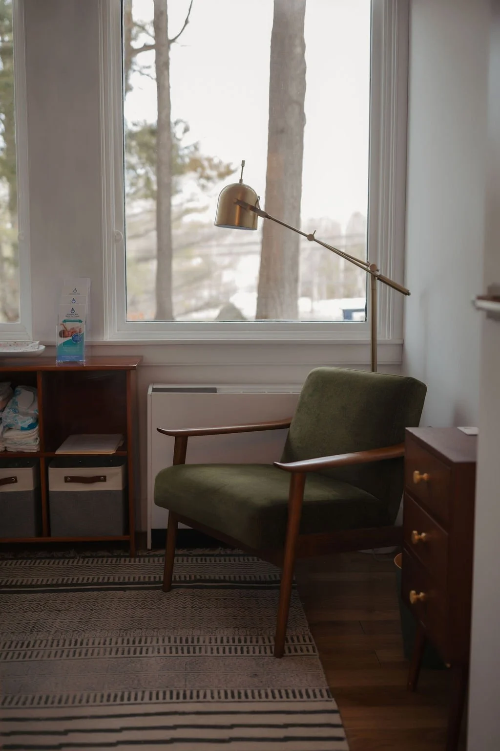 A cozy room with a large window, a green armchair, a wooden side table with drawers, a modern brass reading lamp, and a small wooden cabinet, with trees visible outside the window.
