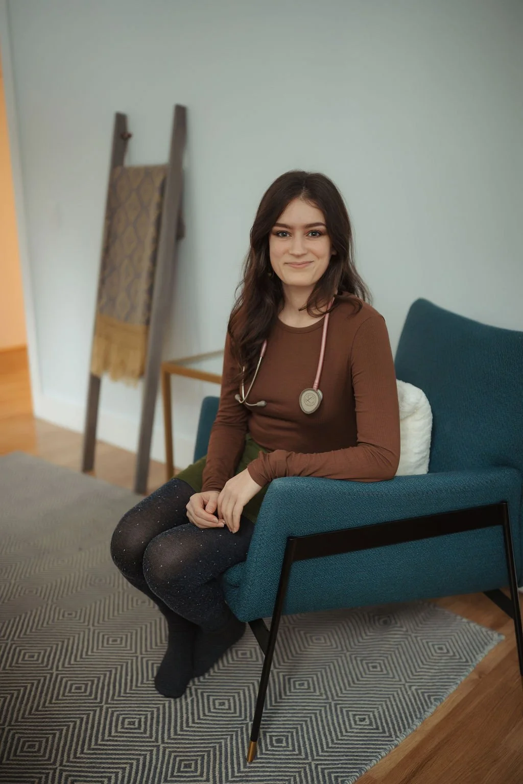 A young woman with long brown hair wearing a brown long-sleeve shirt and black sparkly tights, sitting on a blue armchair with a stethoscope around her neck, in a room with a patterned rug and an easel in the background.