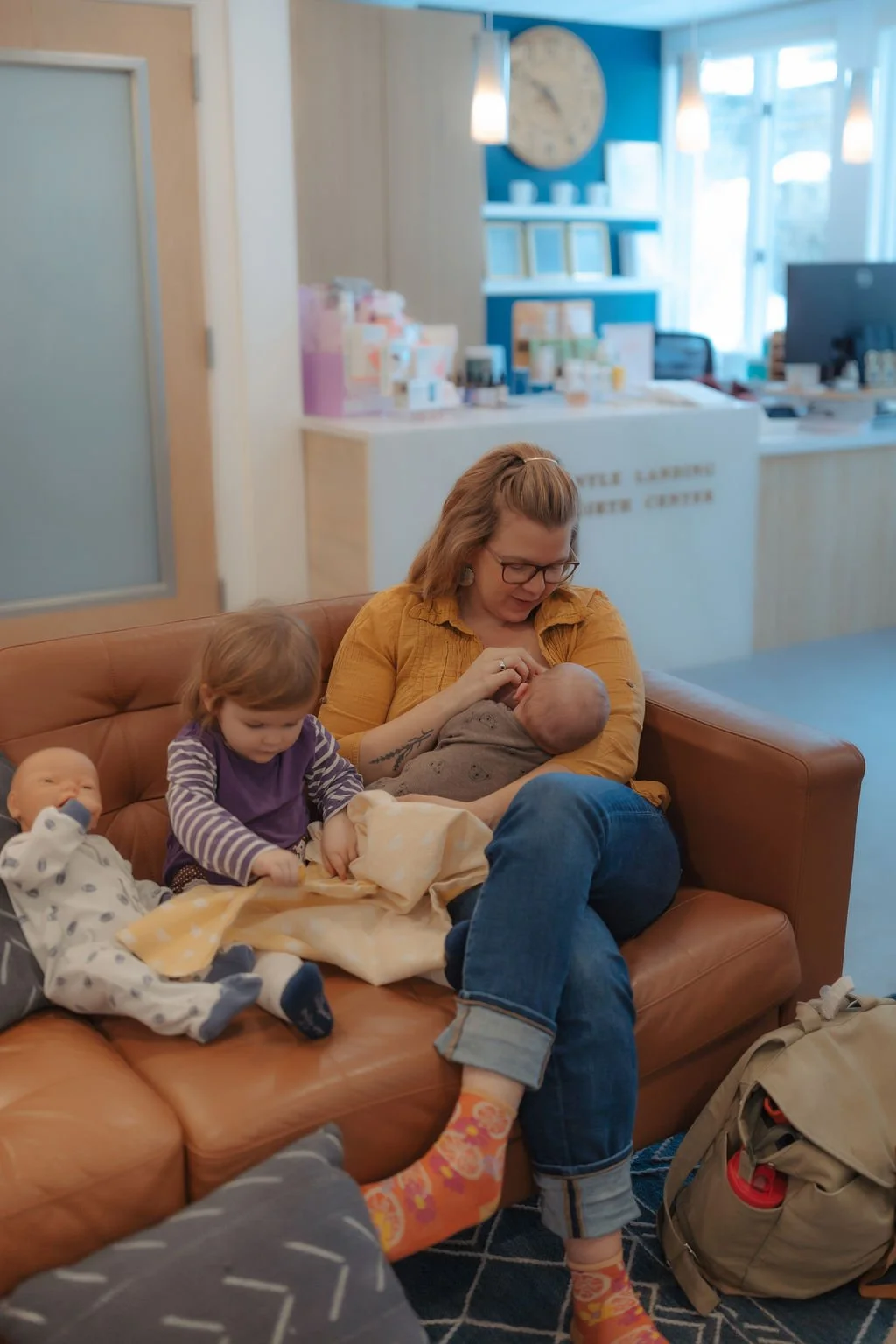 A woman breastfeeding a baby on a tan couch while two young children sit nearby in a waiting or reception area.
