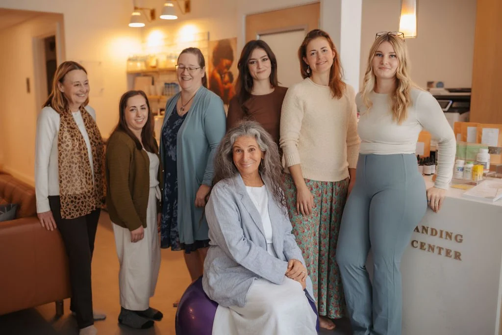 Seven women posing together indoors, one seated on a purple exercise ball, others standing behind, in a well-lit room with warm decor and a table on the right labeled 'Branding Center'.