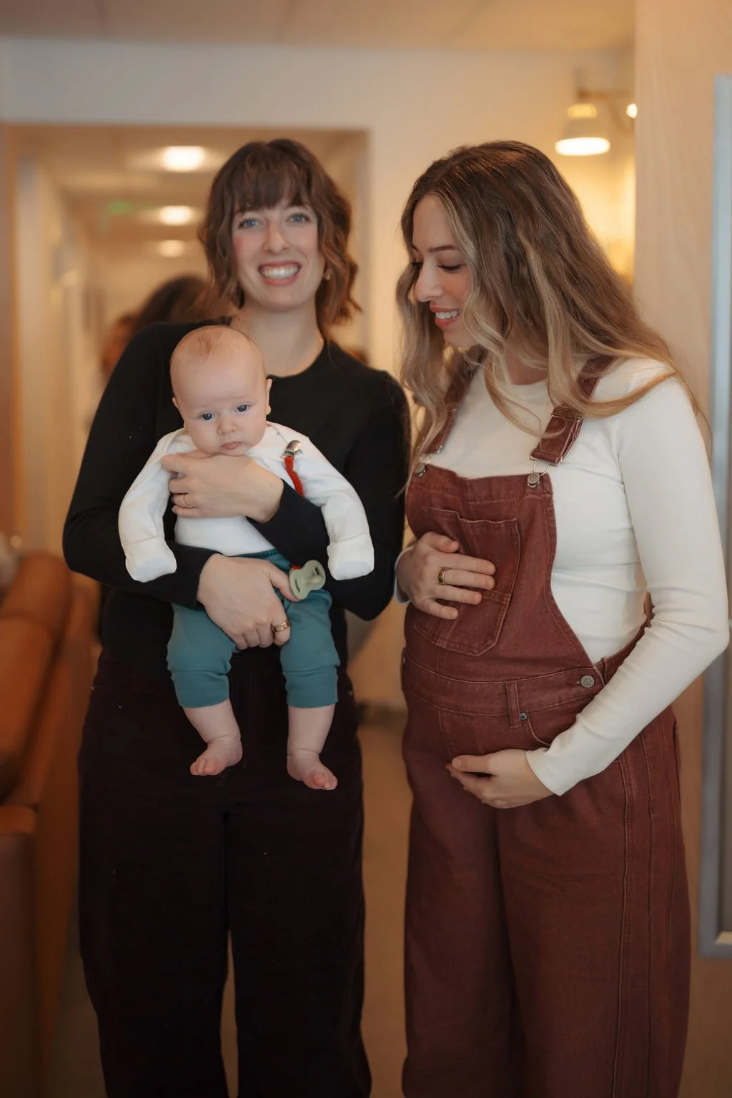 Two women standing indoors, one holding a baby, smiling and looking at the baby, with warm lighting and a blurred background.