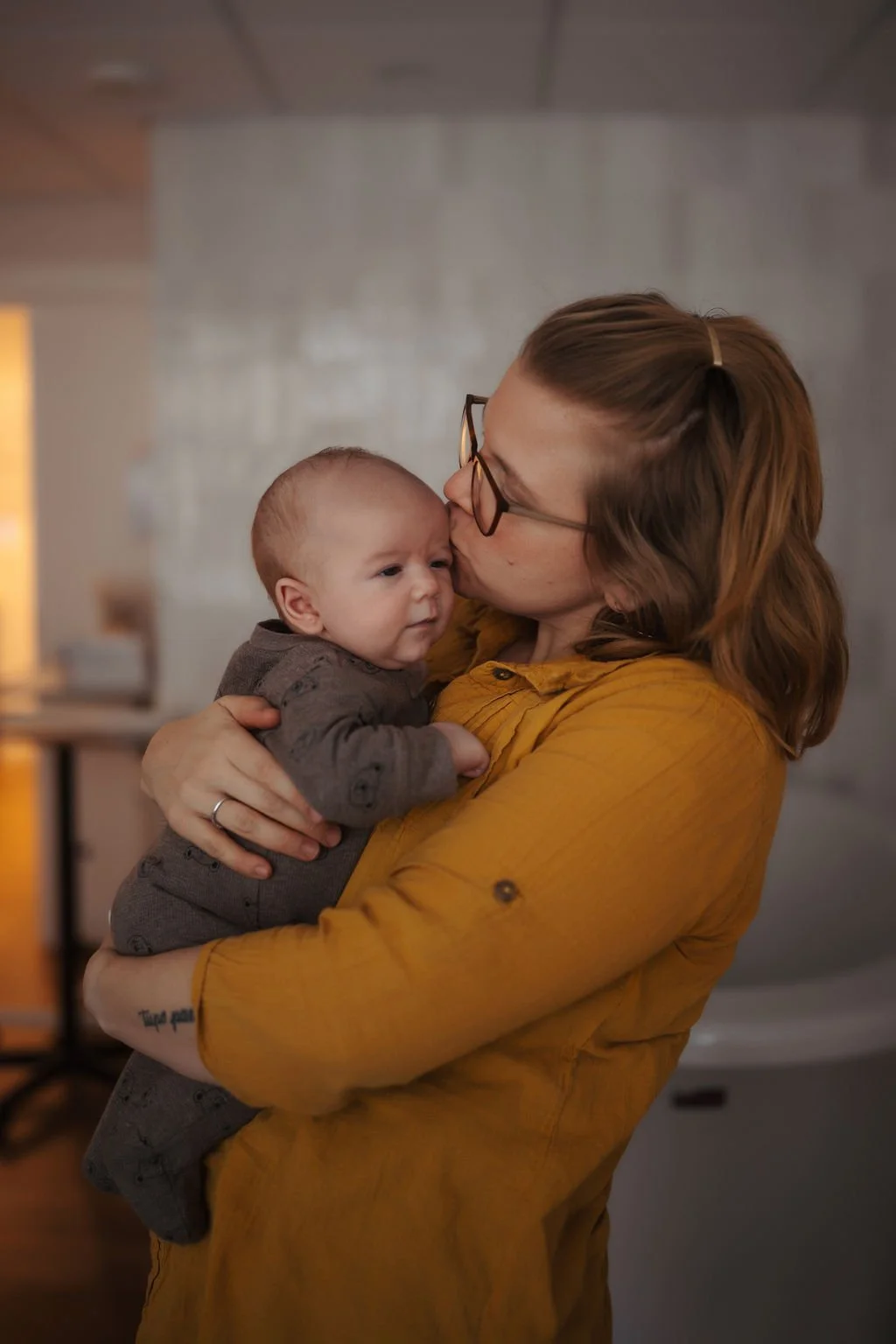 A woman with glasses and shoulder-length hair, wearing a mustard yellow shirt, is holding and kissing a baby on the forehead inside a home.
