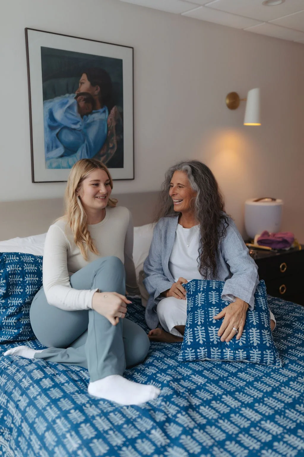 A young woman and an older woman sit on a bed, smiling and talking in a well-lit room with framed art on the wall behind them.