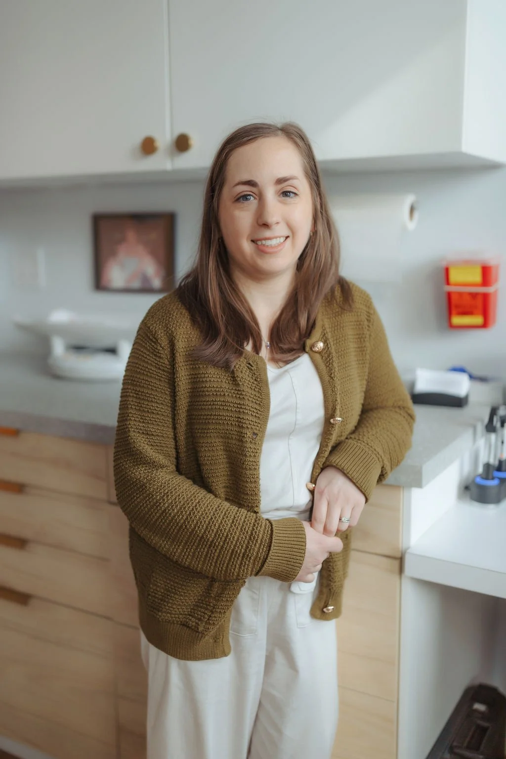 A woman standing in a room with a smile, wearing a brown cardigan and white pants, with a white top underneath. Behind her are kitchen cabinets, a framed picture, and some items on the countertop.