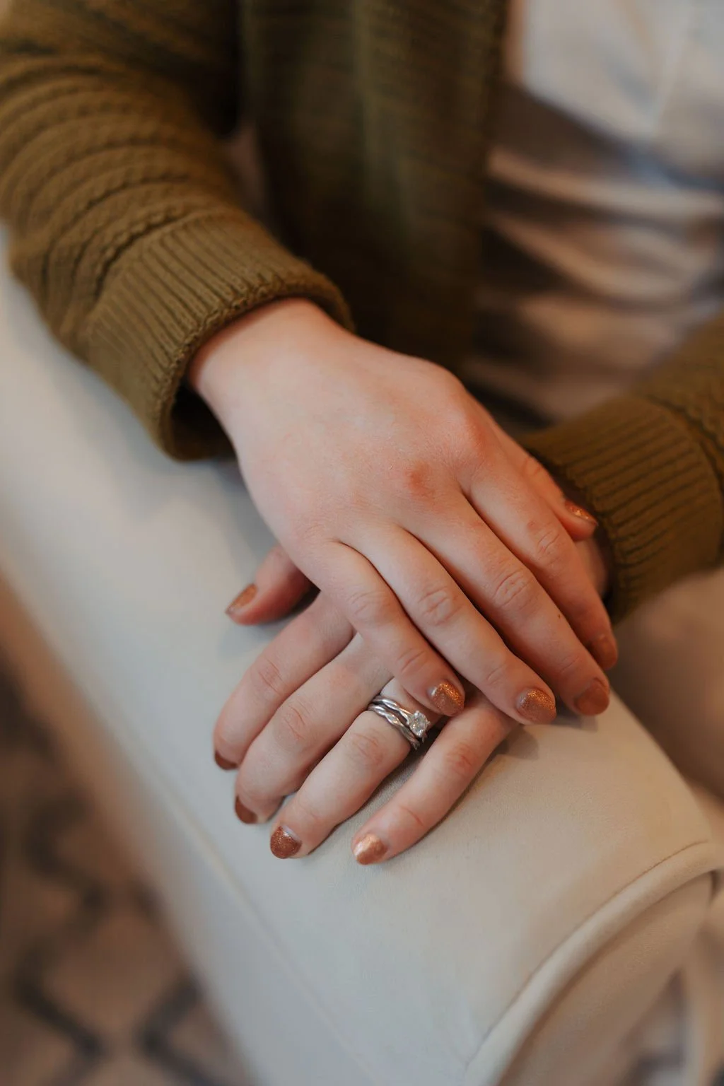 Close-up of two hands with wedding rings resting on a cream-colored armrest, both wearing sweaters, one in brown and one in beige.