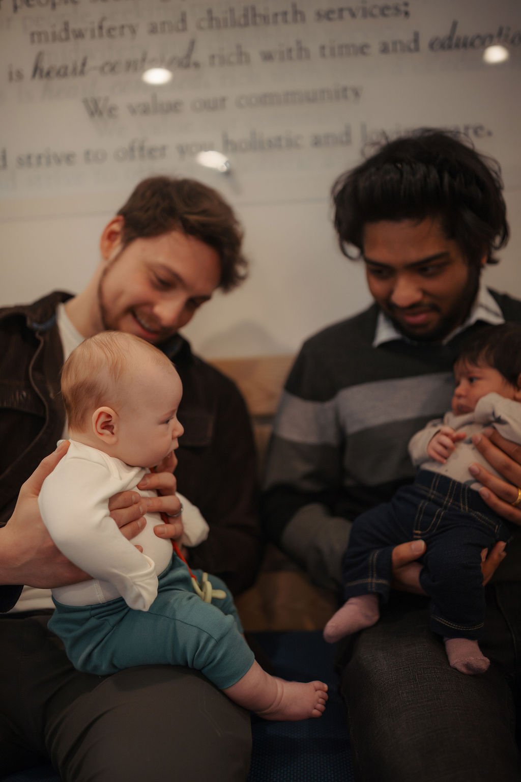 Two men each holding a baby, looking at the babies, with a blurred background that includes text on a wall.