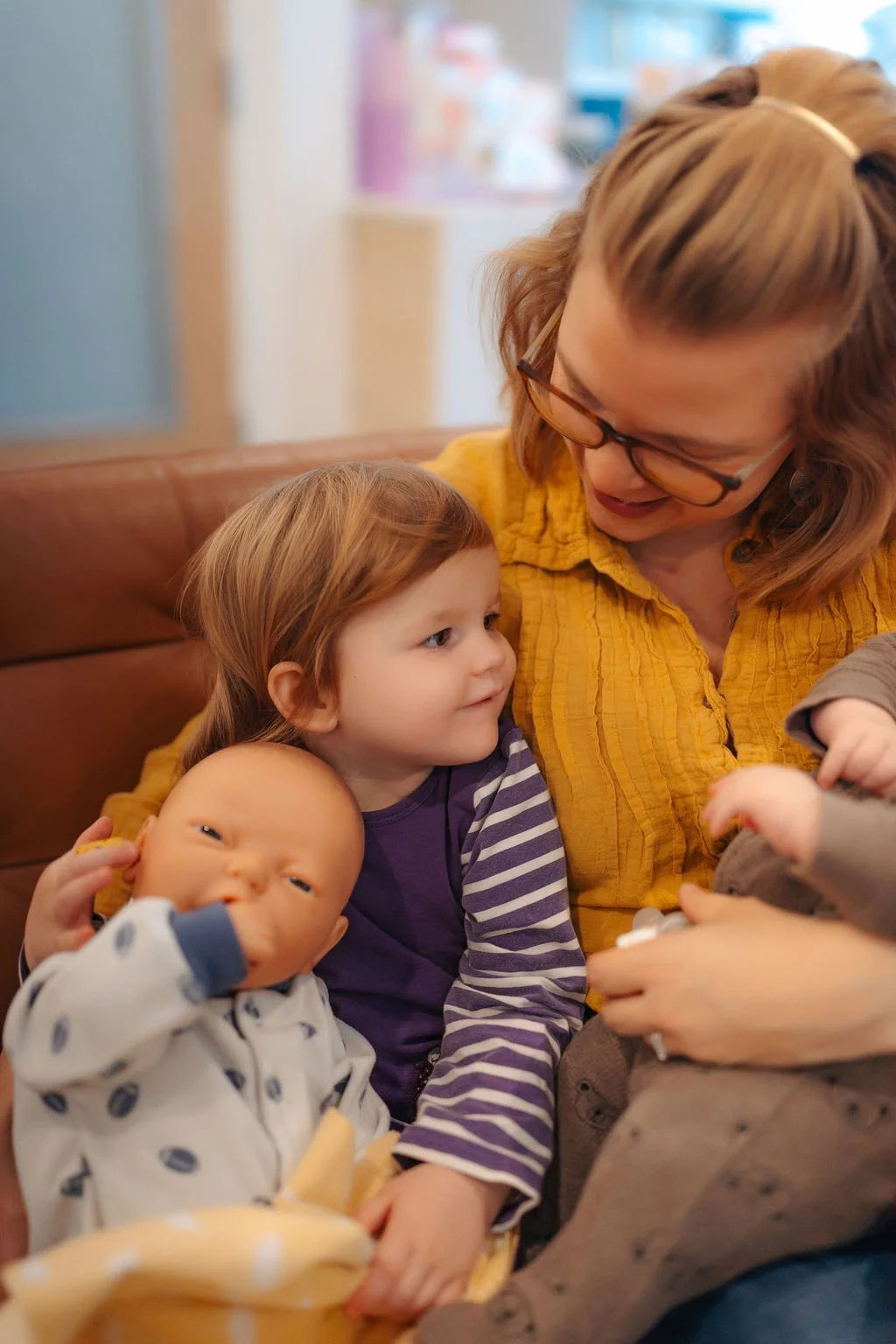 A mother with glasses and two young children, one toddler and one infant, sitting together on a brown couch. The mother is smiling and looking at the toddler, who is holding a bottle. The toddler is looking at the mother, and the infant is drinking f
