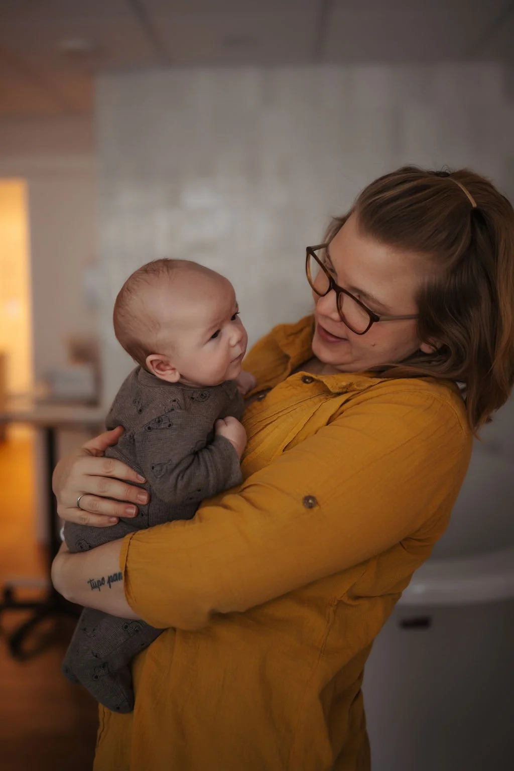 A woman with red hair, glasses, and wearing a mustard yellow shirt, holding a baby and smiling at the baby indoors.