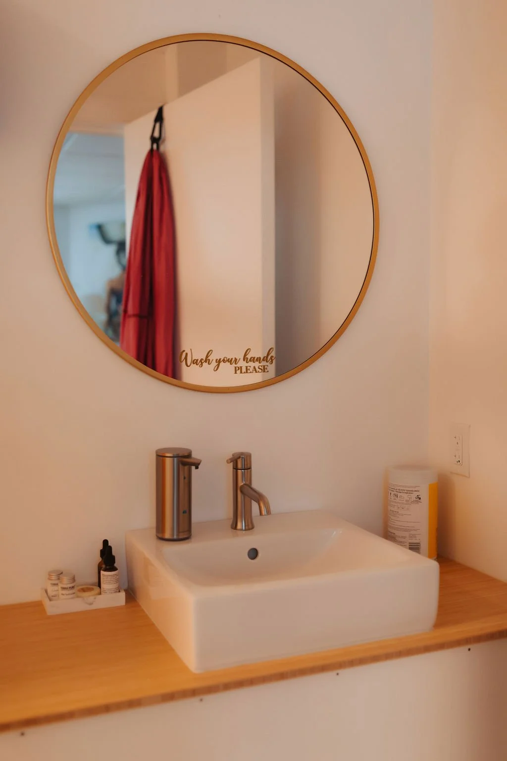 Round wall mirror with a golden frame reflecting a white wall and a red curtain. Text on mirror says "Wash your hands PLEASE." Below the mirror is a white rectangular sink with a modern silver faucet on a wooden countertop. To the left of the sink, s