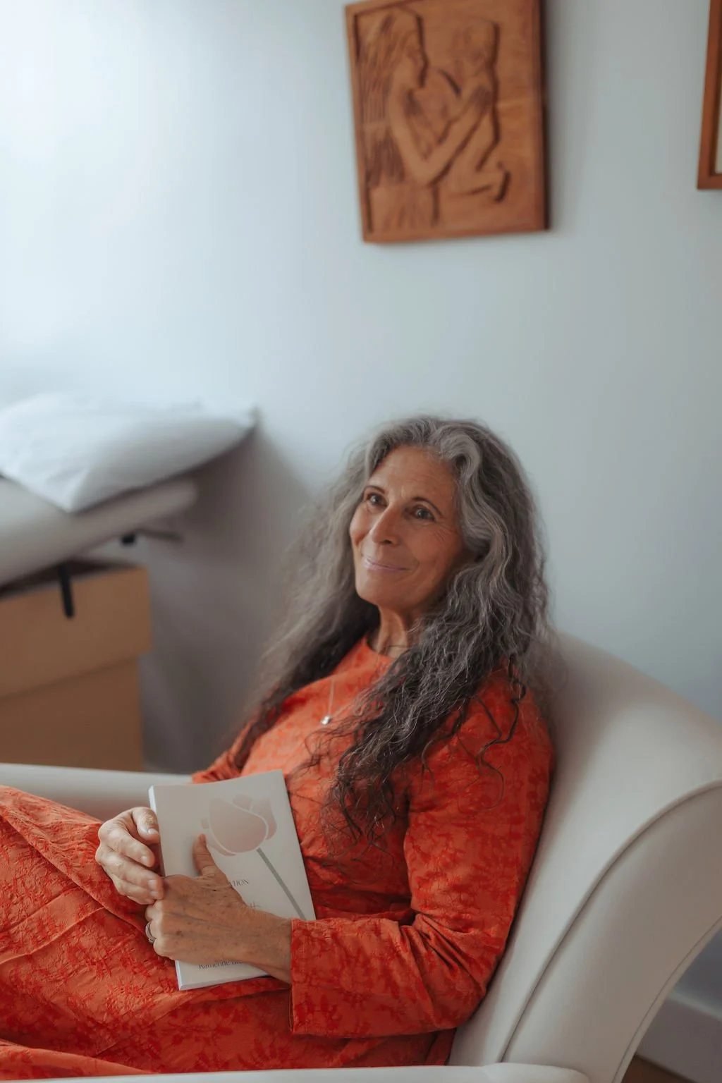 An elderly woman with long gray hair sitting on a beige chair, holding a book, and smiling gently. She is wearing an orange dress. The background includes a white wall and a wooden frame with an abstract artwork.