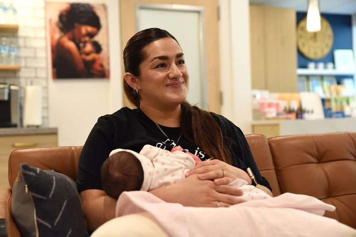 A woman with dark brown hair sitting on a brown leather couch, holding a newborn baby wrapped in a pink blanket, smiling gently. The background shows a cozy indoor setting with shelves, artwork, and warm lighting.