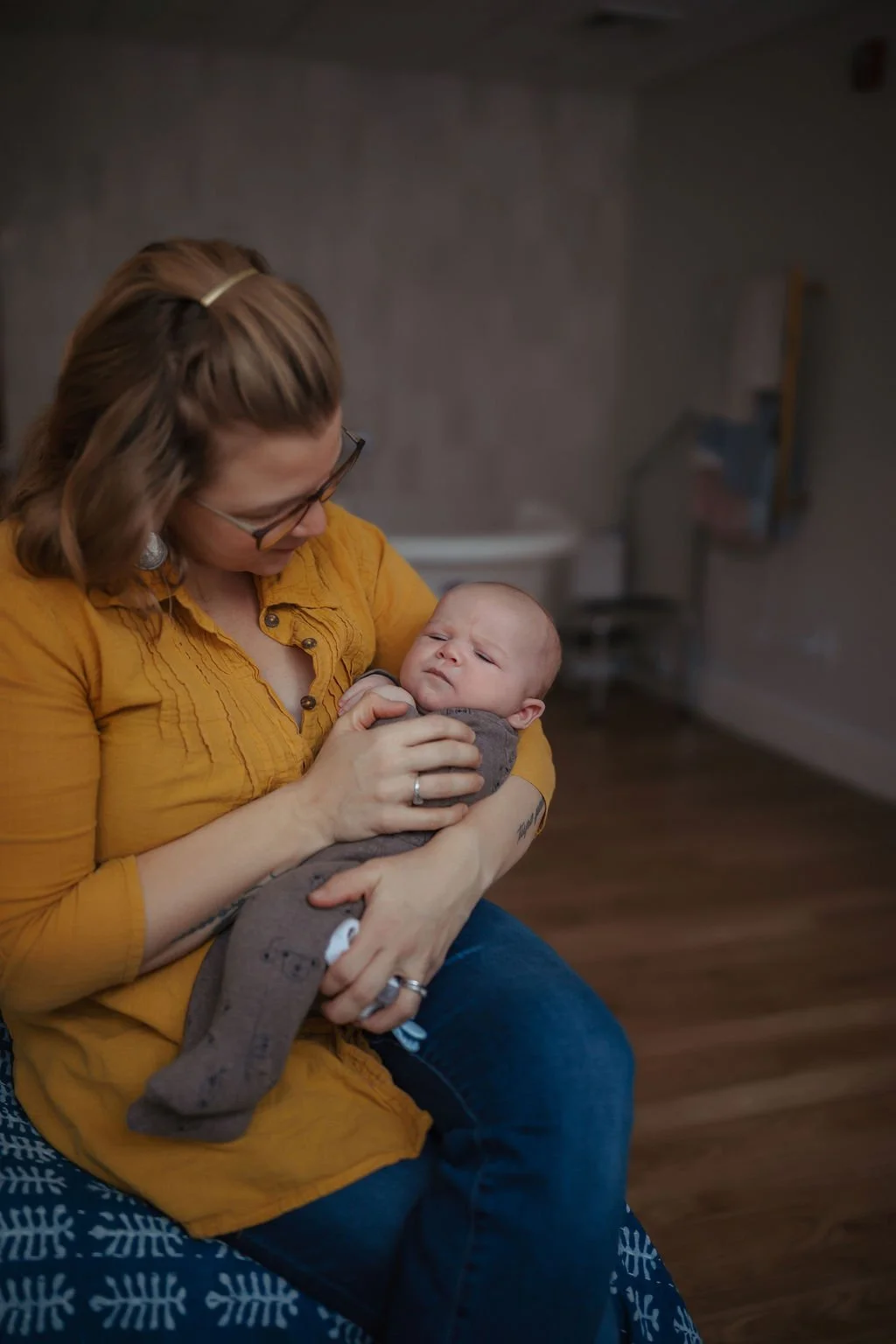 A woman with glasses and a mustard yellow shirt sitting on a blue and white patterned surface holding a baby in her arms. The woman is looking down at the baby, who is wearing a brown outfit, and they are in a room with a dark background.
