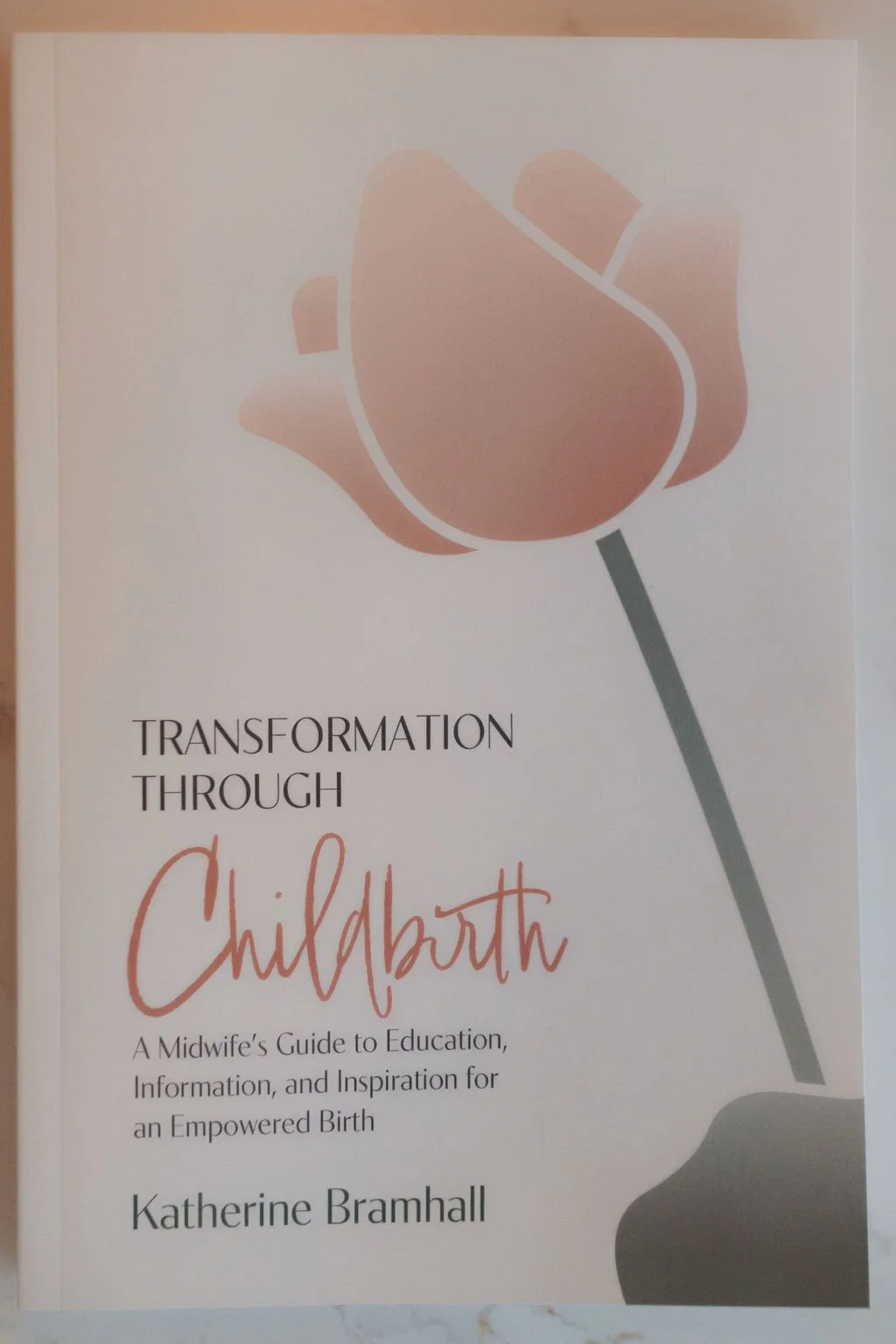 Book cover titled "Transformation Through Childbirth" by Katherine Bramhall, with a stylized illustration of a pink flower.
