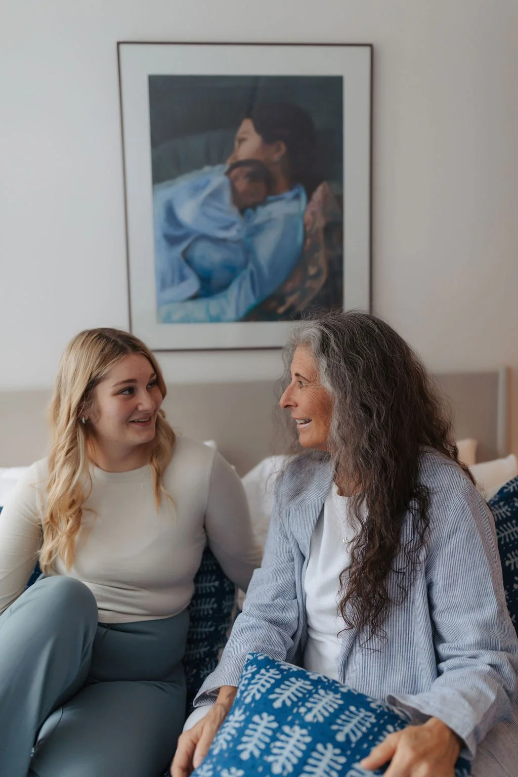A young woman and an older woman are sitting on a bed, smiling and talking to each other in a bedroom with a painting of a woman holding a dog on the wall behind them.