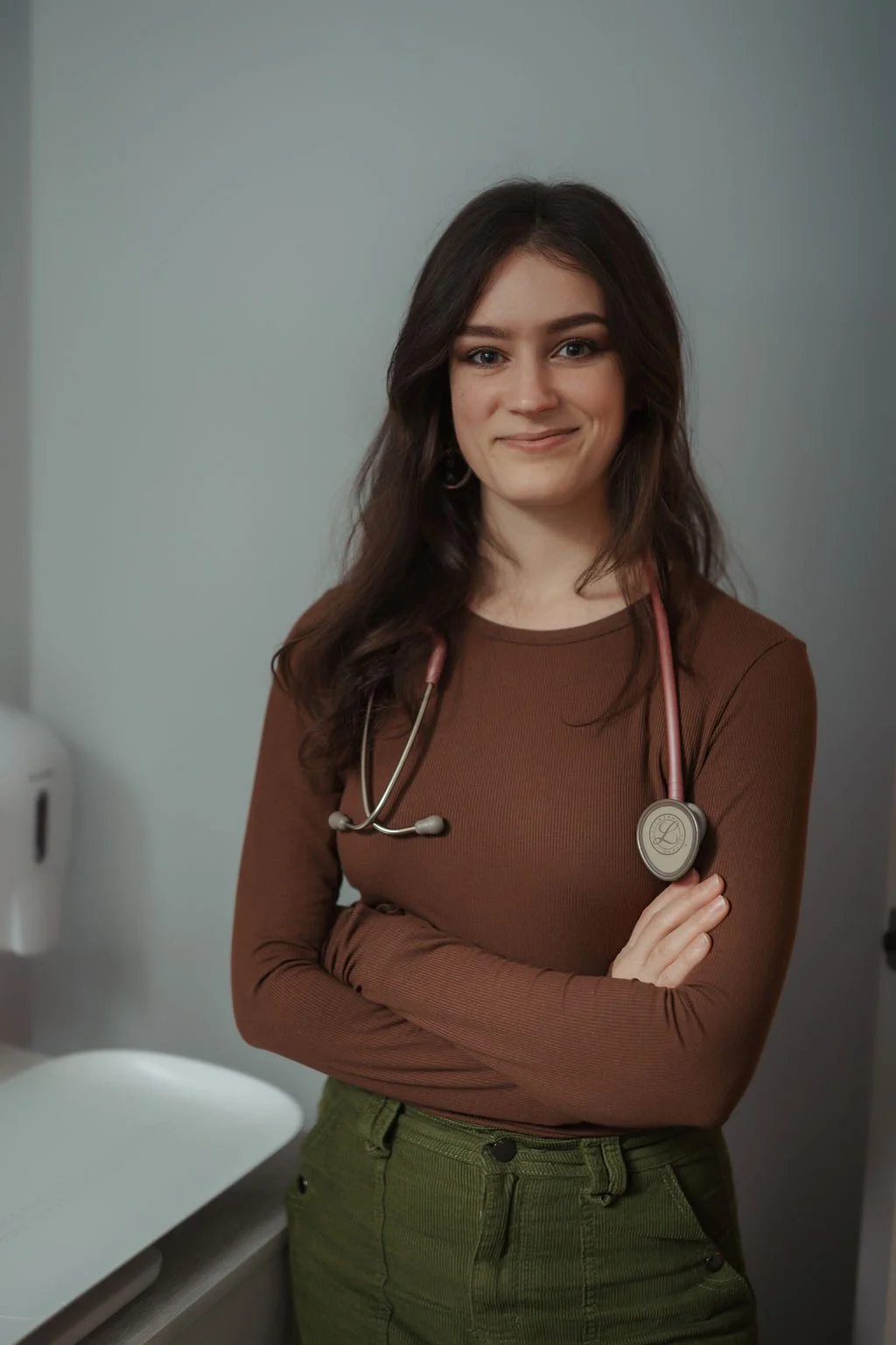 A young female healthcare professional with dark brown hair, wearing a brown long-sleeved top and green pants, smiles confidently at the camera in a medical setting. She has a pink stethoscope around her neck and crossed arms.