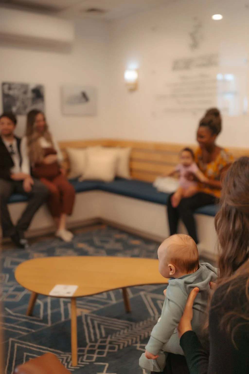 A woman holding a baby, with a blurred background of people sitting on a corner bench in a cozy room, with a patterned carpet and wall art.