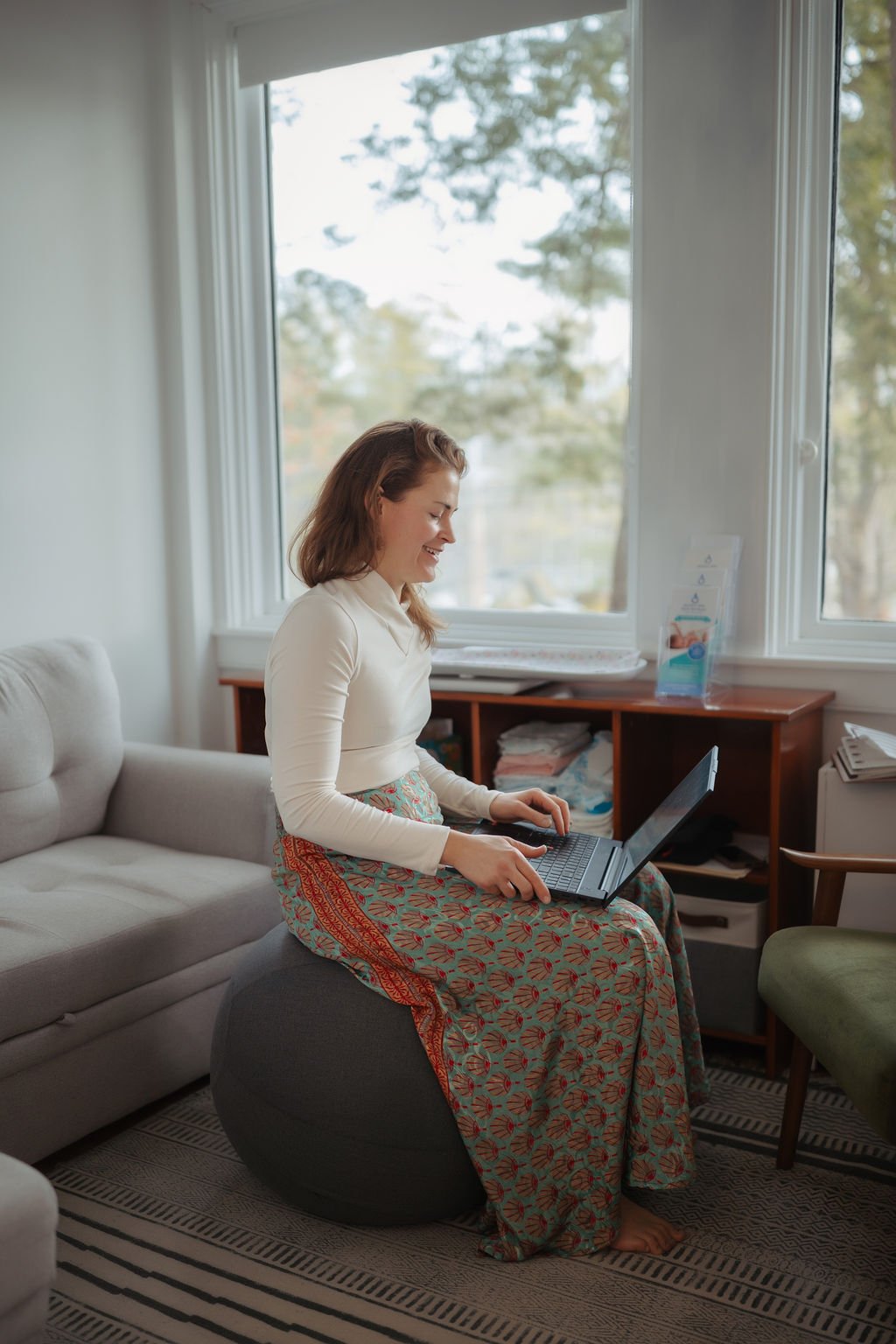 A woman sitting on an exercise ball in a living room, smiling while using a laptop. She is wearing a long, patterned skirt and a white long sleeves shirt. The room has a large window with a view of trees outside, and various books and papers on a woo