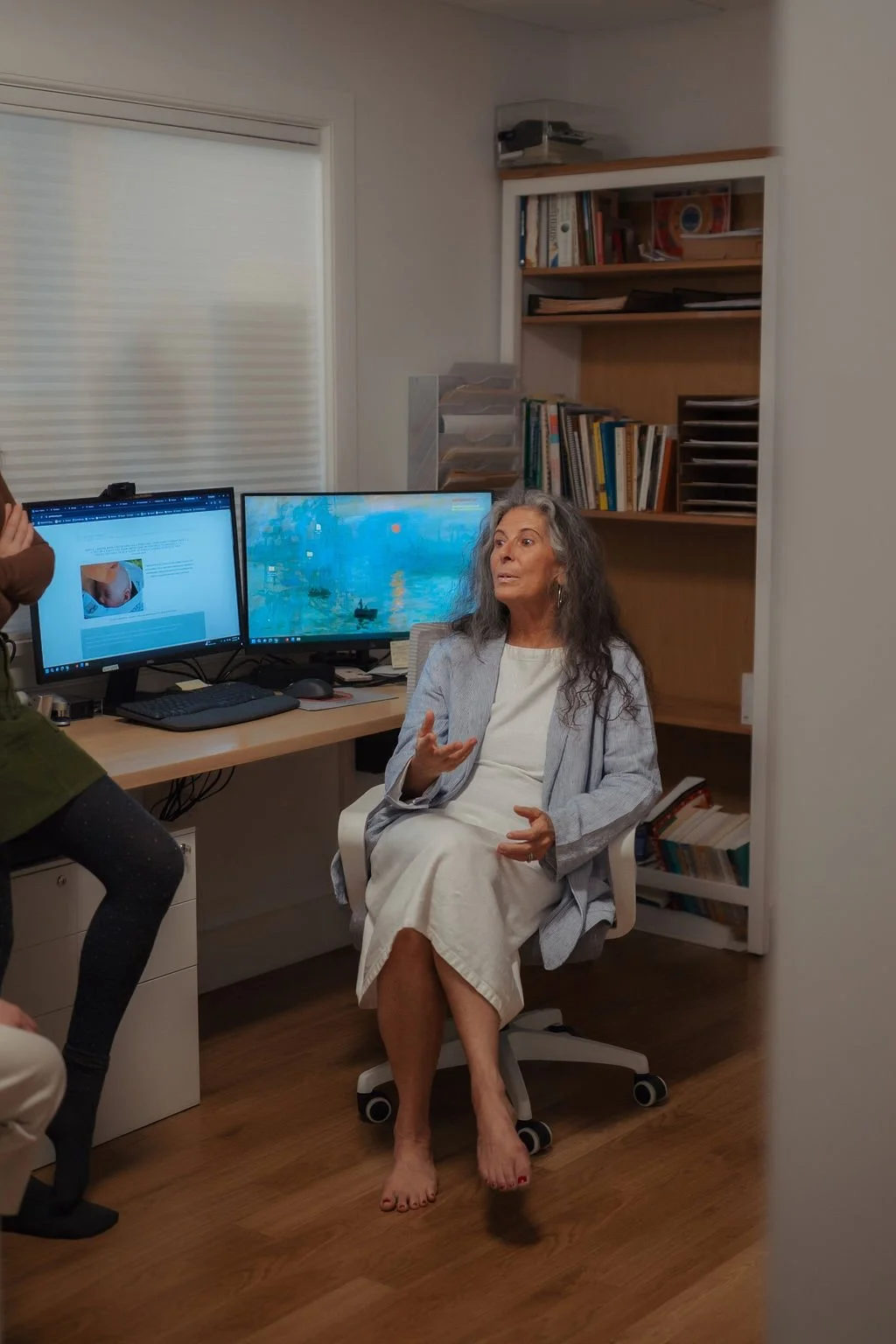 An elderly woman with gray hair seated in a white office chair, speaking and gesturing with her hands. She is in a home office with two computer monitors on a desk, paperwork on a bookshelf in the background, and another person standing nearby.