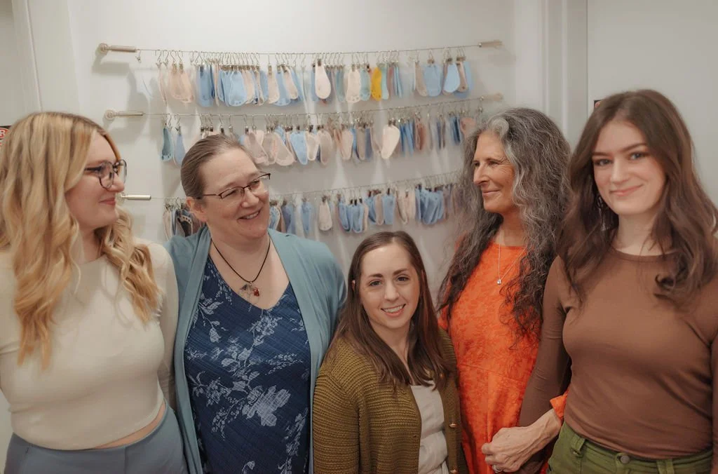 Five women standing together inside a room with a display of baby shoes hanging on the wall behind them, all smiling and talking.