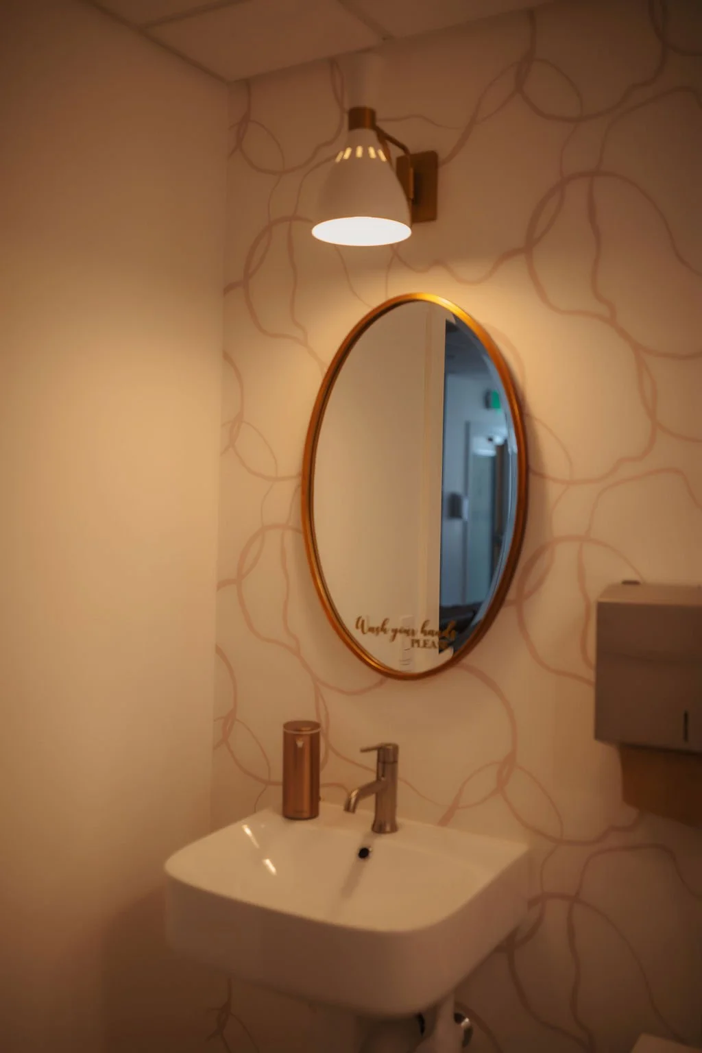A bathroom corner with a wall-mounted mirror, a white sink, and a wall-mounted paper dispenser. The wall behind has a light pattern with interconnected lines, and a wall sconce with a white shade provides warm lighting.